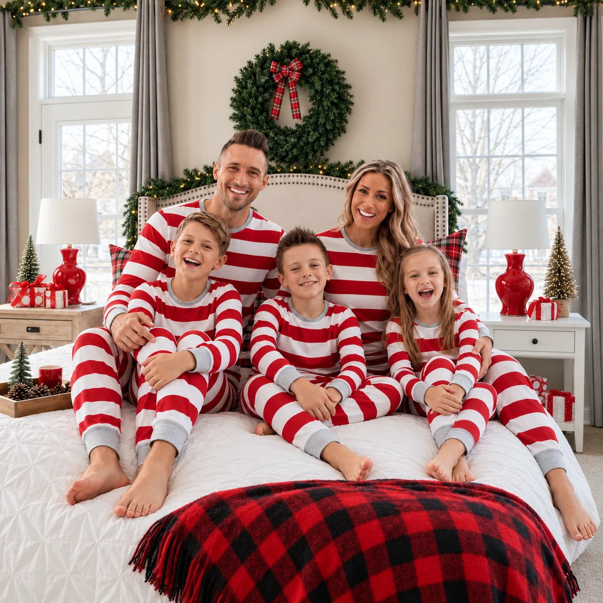 Family in red and white striped Christmas pajamas sitting on a bed with a red plaid blanket beneath a wreath.