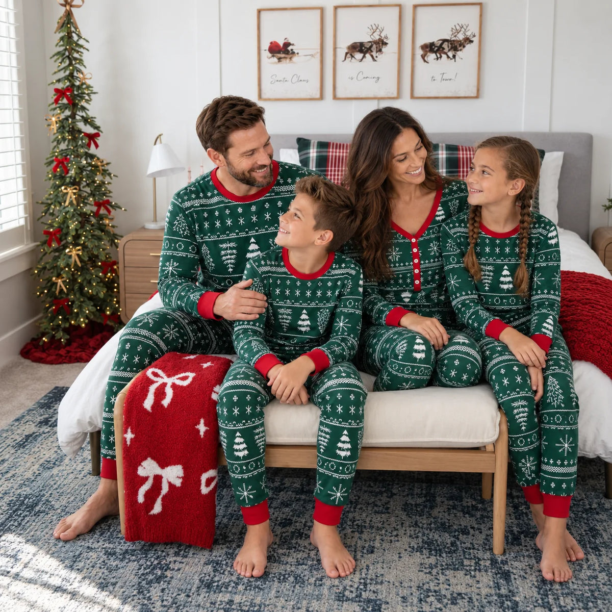 Family in dark green Fair Isle Christmas pajamas sitting on a chair beside a bed, small tree and red blanket.