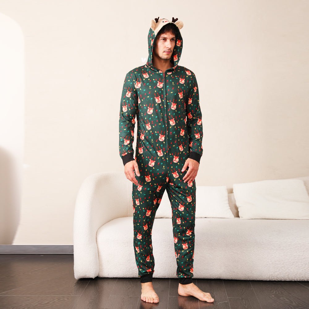 Man modeling a green Christmas print zip-up onesie with a reindeer face hood, antlers and black cuffs beside a sofa.