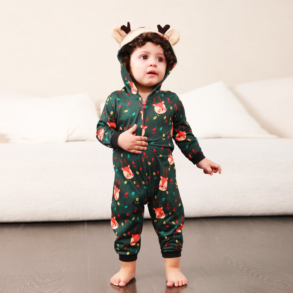 Toddler standing beside a sofa in a green Christmas print zip-up onesie with a reindeer face hood and antlers.