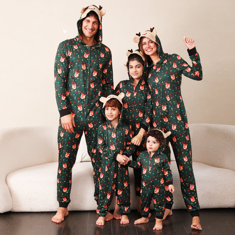 Family standing beside a sofa in green Christmas print onesies with reindeer face hoods, antlers and black cuffs.