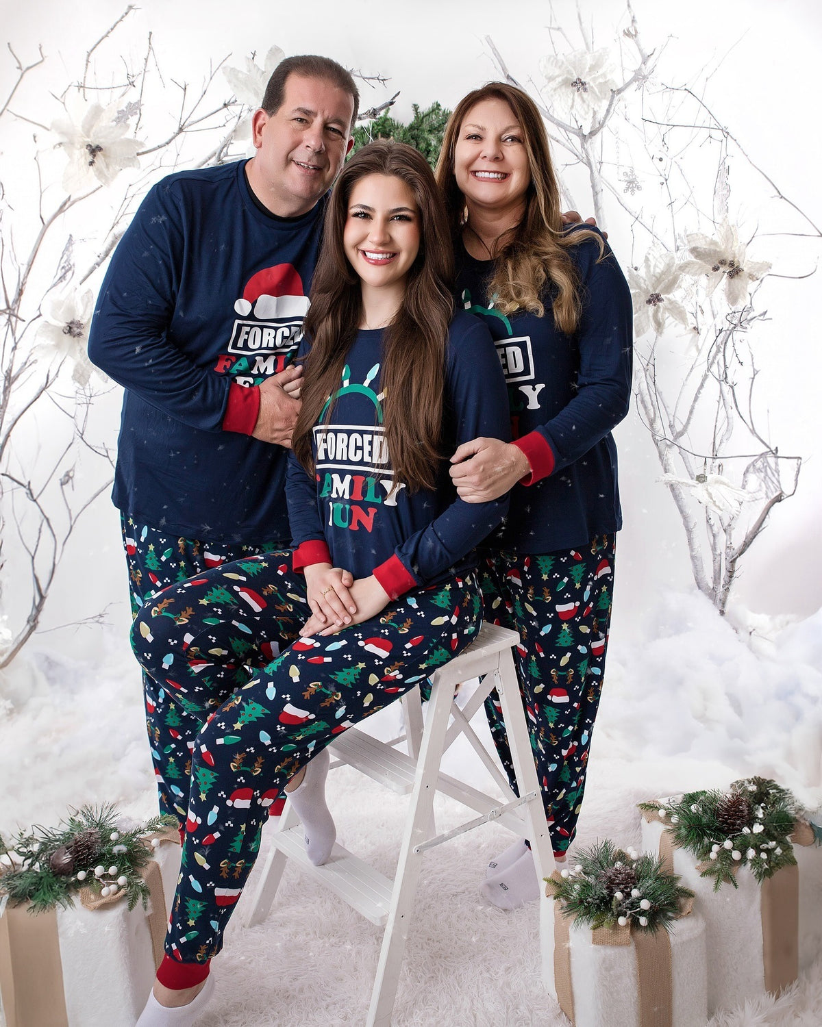 Three adults in navy Forced Family Fun pajamas with red cuffs and Christmas print pants among white holiday branches.