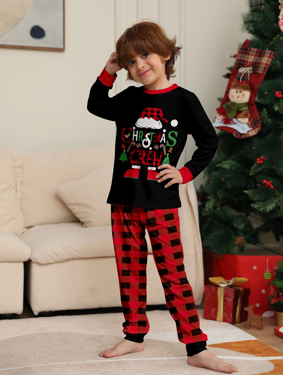 Bambino in posa accanto a un albero di Natale con maglia nera Christmas Crew del pigiama con polsini rossi e pantaloni a grandi quadri.