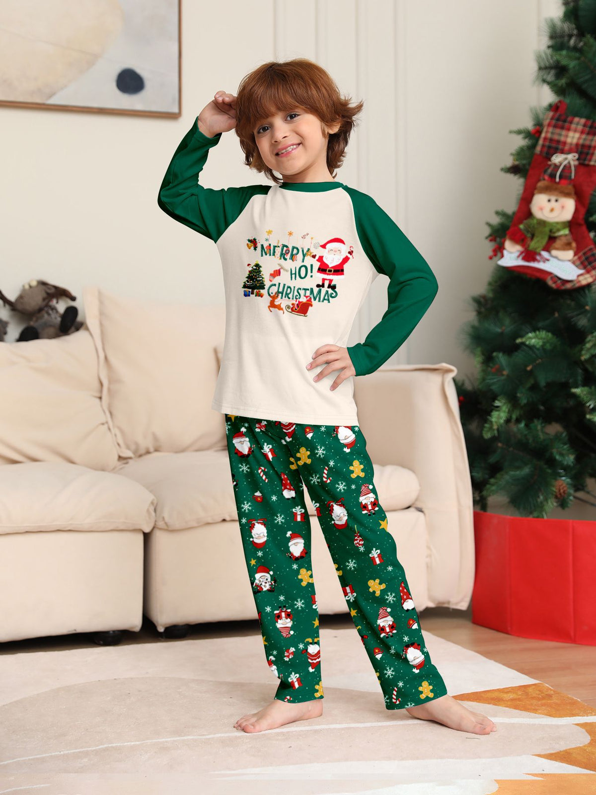 Bambino in posa accanto a un albero con maglia color crema del pigiama con motivo natalizio e maniche verdi e pantaloni verdi con Babbo Natale.
