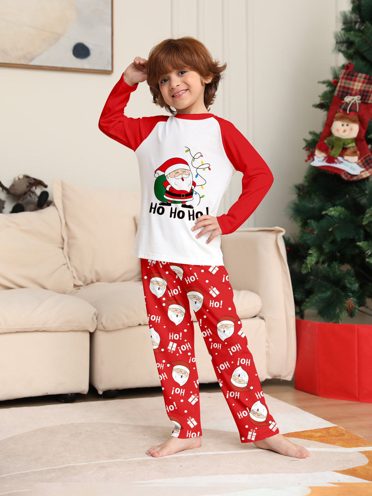 Boy posing beside a Christmas tree in a white Santa pajama top with red sleeves and red Santa print pants.