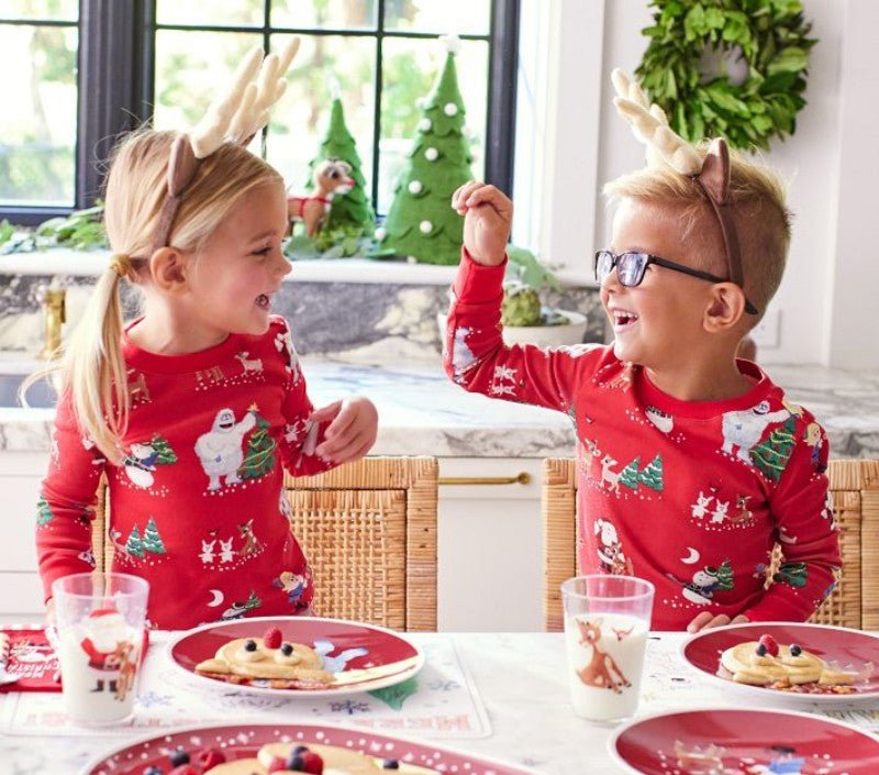 Due bambini con cerchietti con corna che fanno colazione in pigiami natalizi rossi con pupazzi di neve e renne.