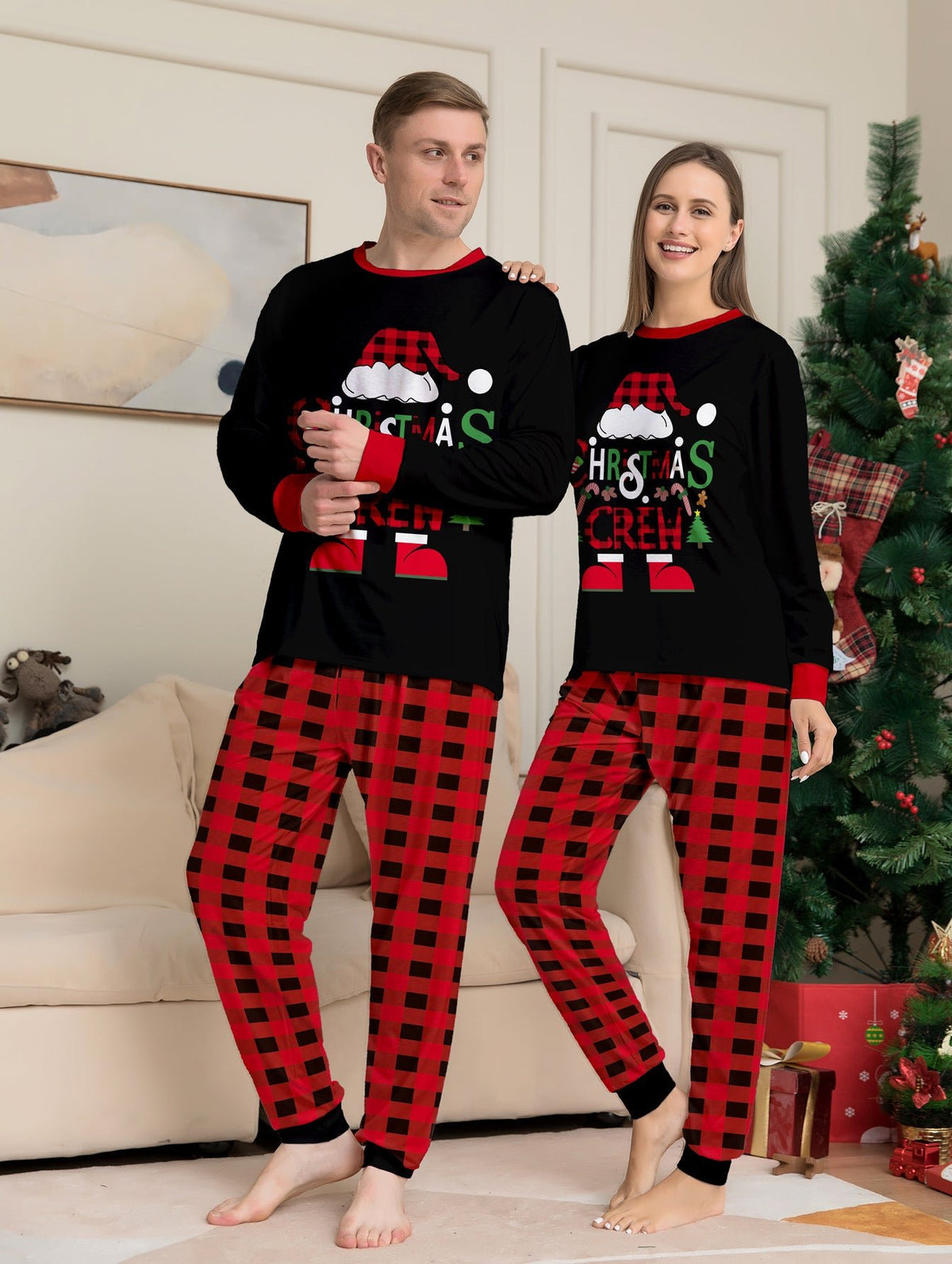 Coppia in posa accanto a un albero di Natale con maglie nere Christmas Crew del pigiama e pantaloni a grandi quadri rossi e neri.