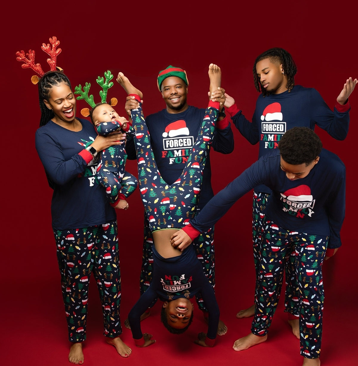 Family holding a child upside down against a red backdrop in navy Forced Family Fun Christmas pajamas with red cuffs.