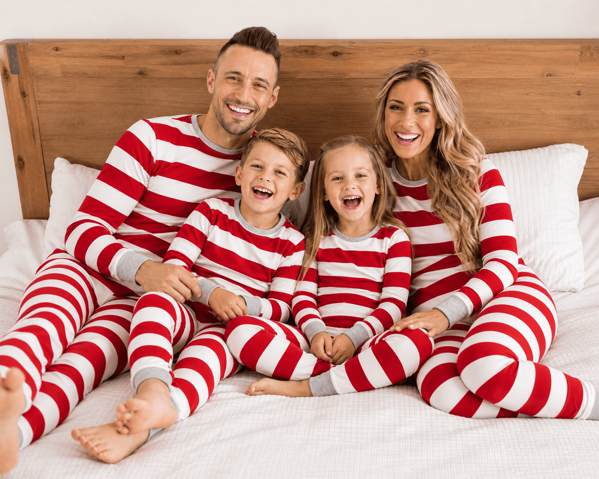 Genitori e due bambini che ridono su un letto, in pigiami natalizi a righe rosse e bianche con polsini grigi.