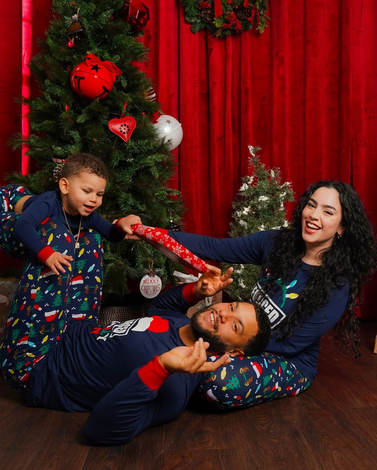 Genitori e un bambino che giocano accanto a un albero in pigiami blu navy Forced Family Fun con pantaloni a stampa natalizia multicolore.