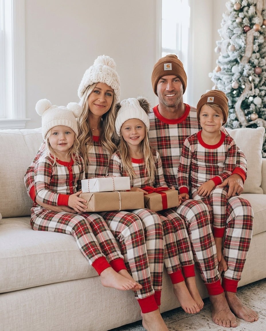 Famiglia con berretti lavorati a maglia e regali incartati in mano su un divano, in pigiami natalizi a quadri beige, rossi e neri.