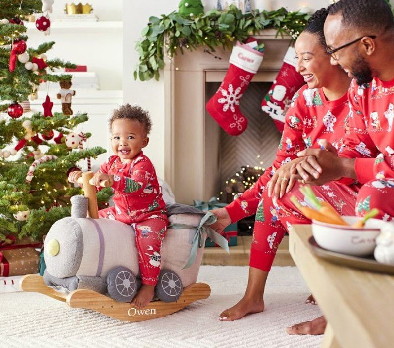 Bambino piccolo su un trenino giocattolo accanto ai genitori e a un albero di Natale, tutti in pigiami rossi con personaggi festivi.