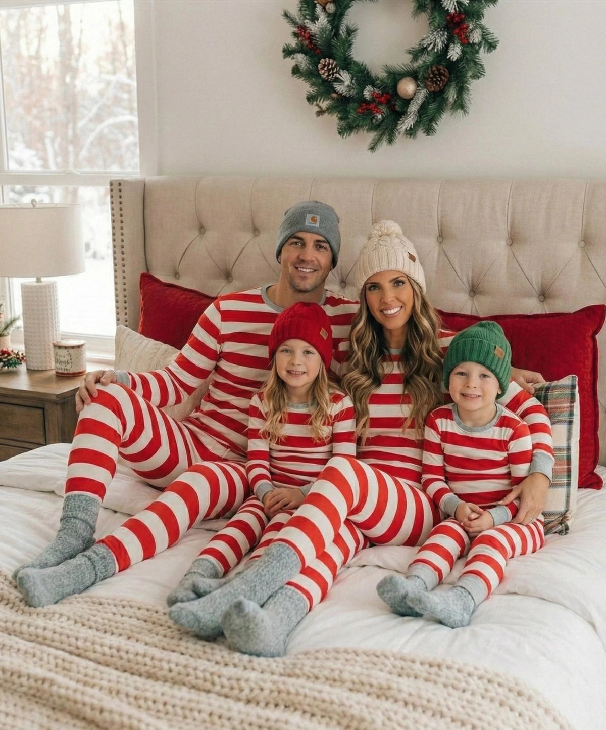 Famiglia che si rilassa su un letto sotto una ghirlanda, in pigiami natalizi a righe rosse e bianche, berretti lavorati a maglia e calzini grigi.