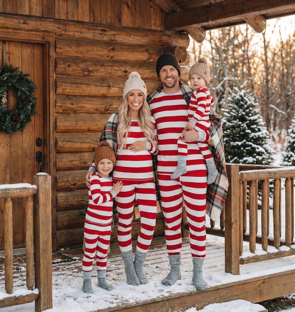 Famiglia con berretti lavorati a maglia e pigiami natalizi a righe rosse e bianche, in piedi sulla veranda innevata di una baita.
