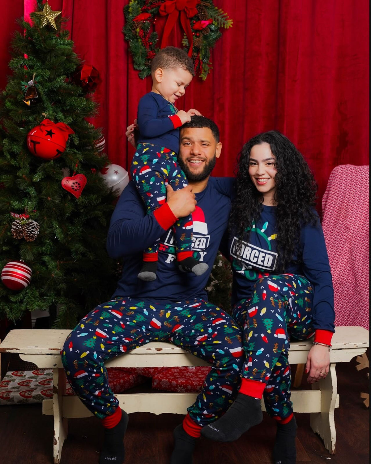 Padre che porta un bambino sulle spalle accanto alla madre, tutti in pigiami blu navy Forced Family Fun accanto a un albero di Natale.