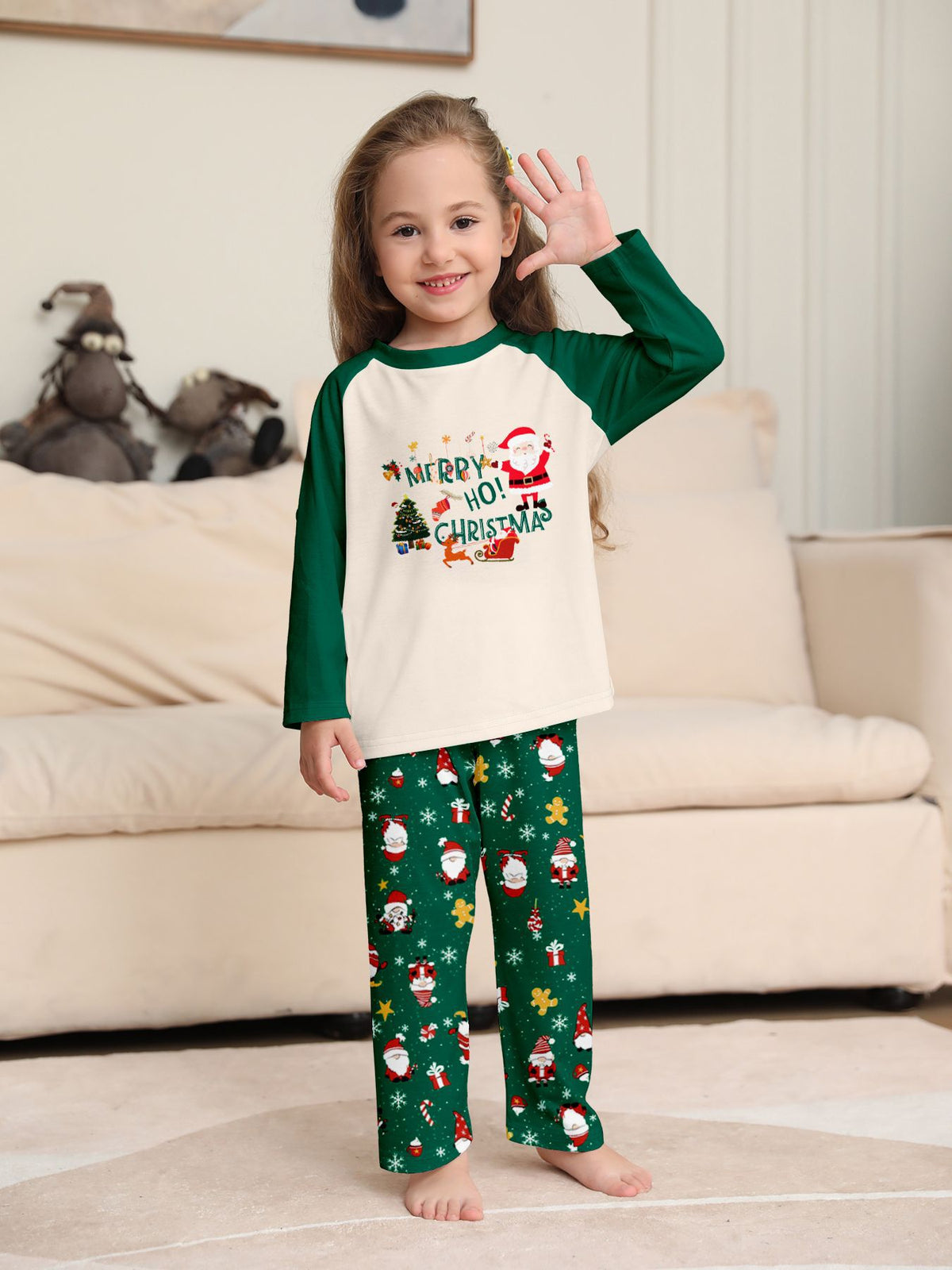 Bambina che saluta con la mano, con maglia color crema del pigiama con motivo natalizio e maniche raglan verdi e pantaloni verdi con Babbo Natale.