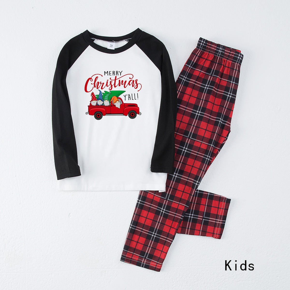 Pigiama per bambini disposto in piano, con maglia bianca Merry Christmas Y'all con camion, maniche raglan nere e pantaloni in tartan rosso.