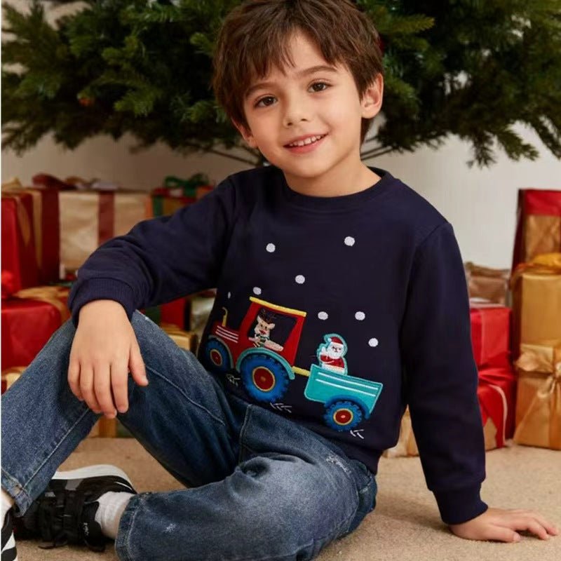 Junge sitzt neben einem Weihnachtsbaum in einem dunkelblauen Sweatshirt mit Rentier auf einem roten Traktor und einem Schneemann im Anhänger.