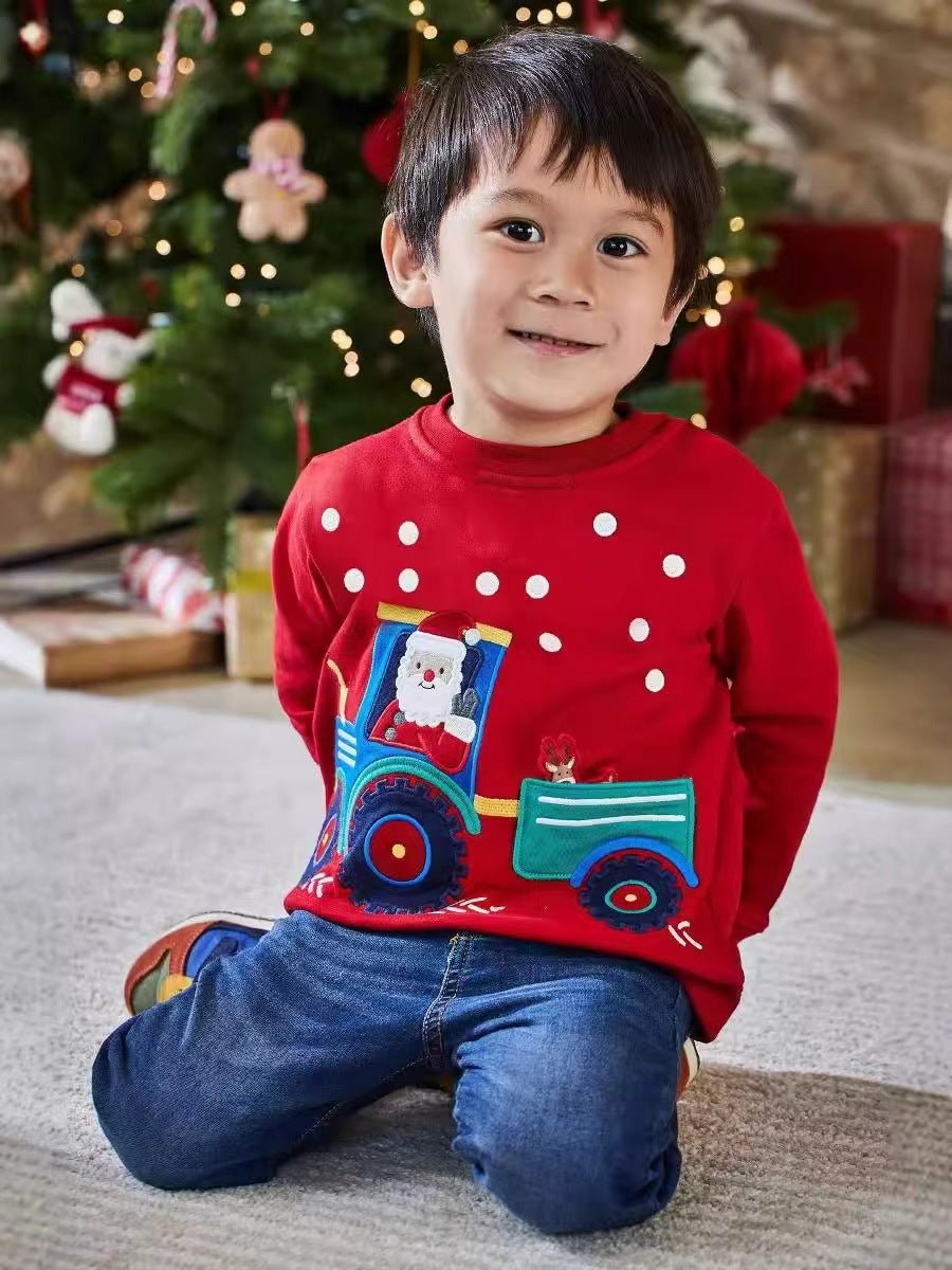 Junge sitzt neben einem Weihnachtsbaum in einem roten Sweatshirt mit Weihnachtsmann auf einem blauen Traktor und einem Rentier im Anhänger.