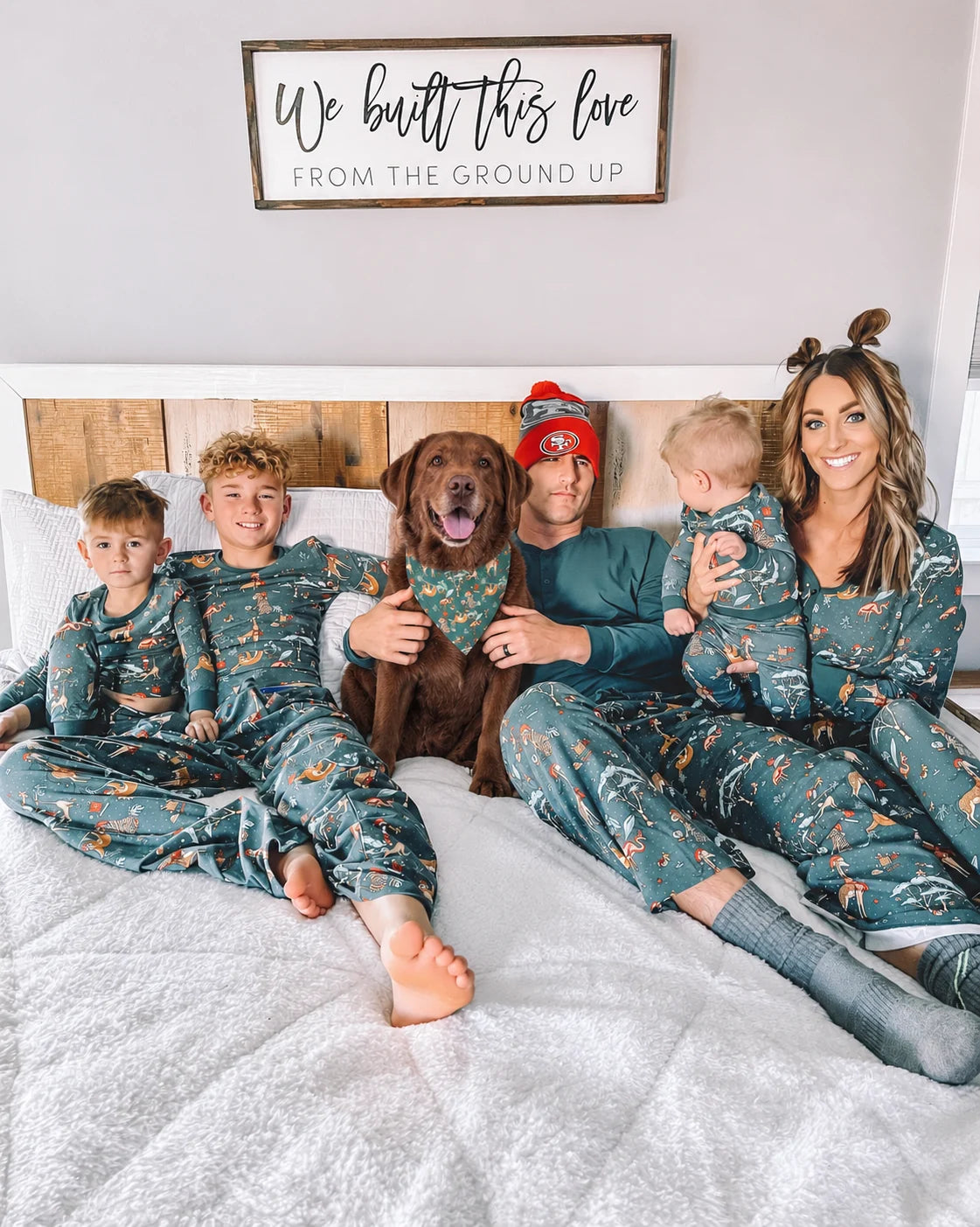 Family in green animal print Christmas pajamas on a bed, with a dog wearing a matching green bandana.