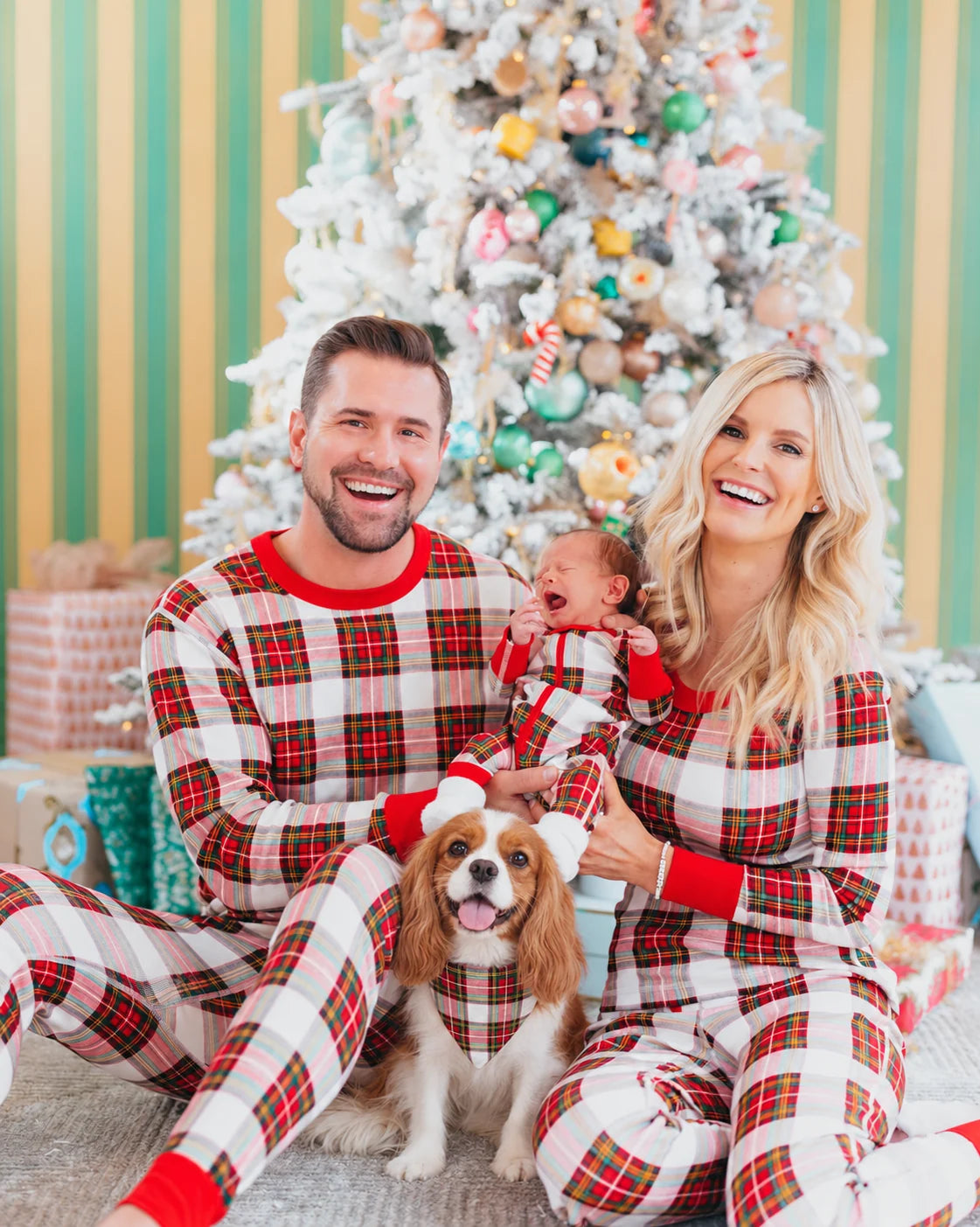 Parents and baby in red, white and black tartan Christmas pajamas beside a dog wearing a matching plaid bandana.