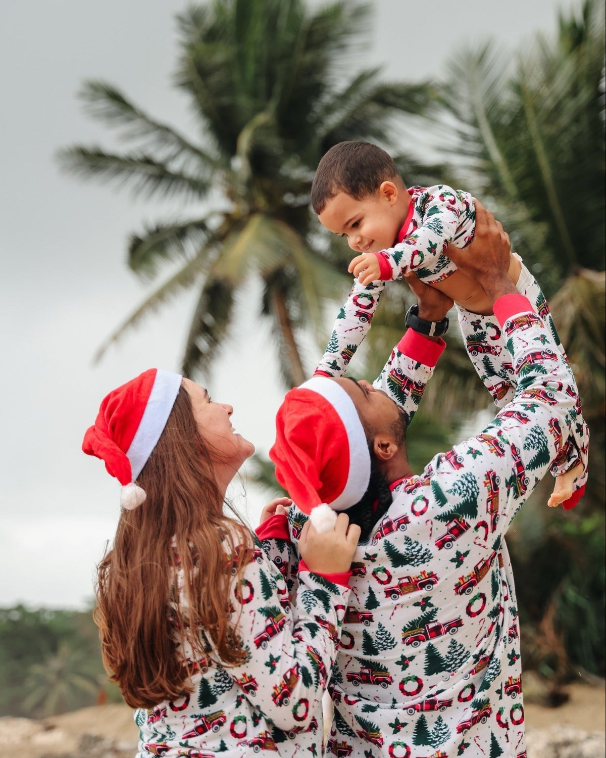 Parents in Santa hats lifting a baby outdoors, all wearing white festive print Christmas pajamas with red cuffs.
