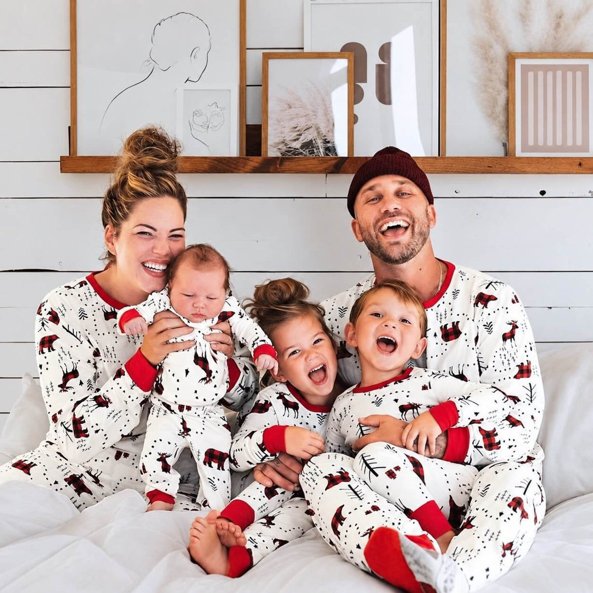 Family of five laughing on a bed in white woodland-print pajamas with red cuffs, including a matching baby outfit.
