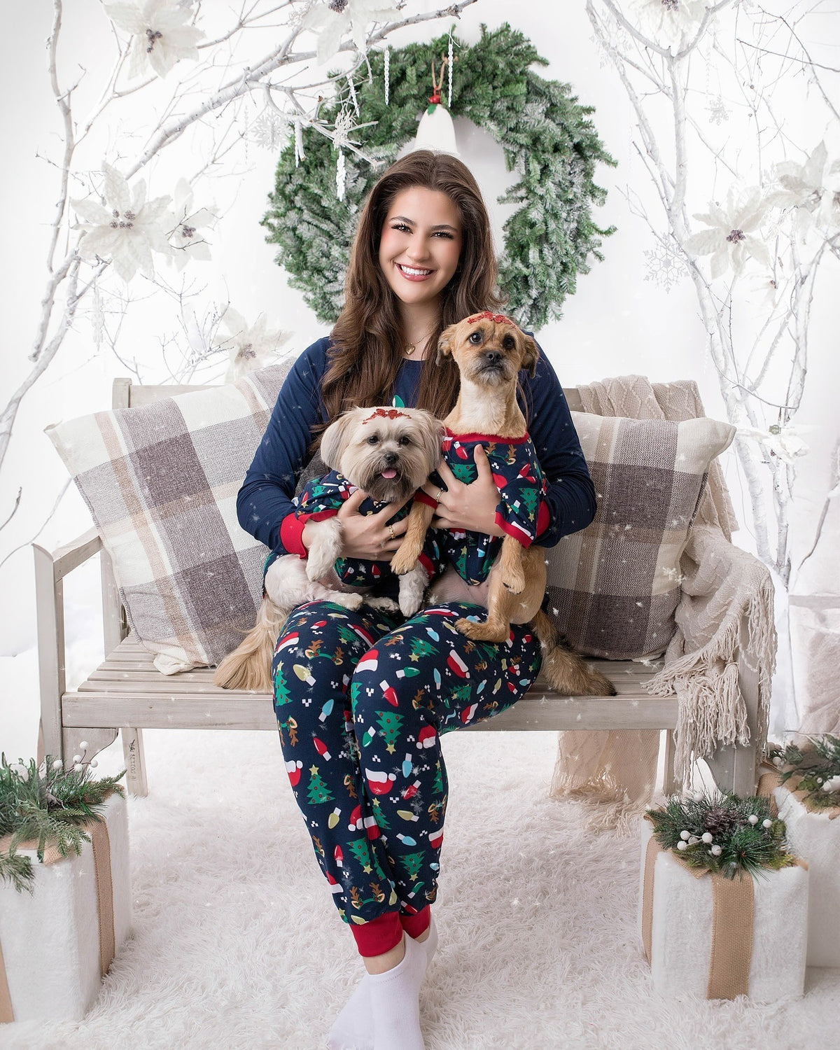 Woman in navy Forced Family Fun pajamas holding two dogs in matching festive outfits on a bench among white branches.