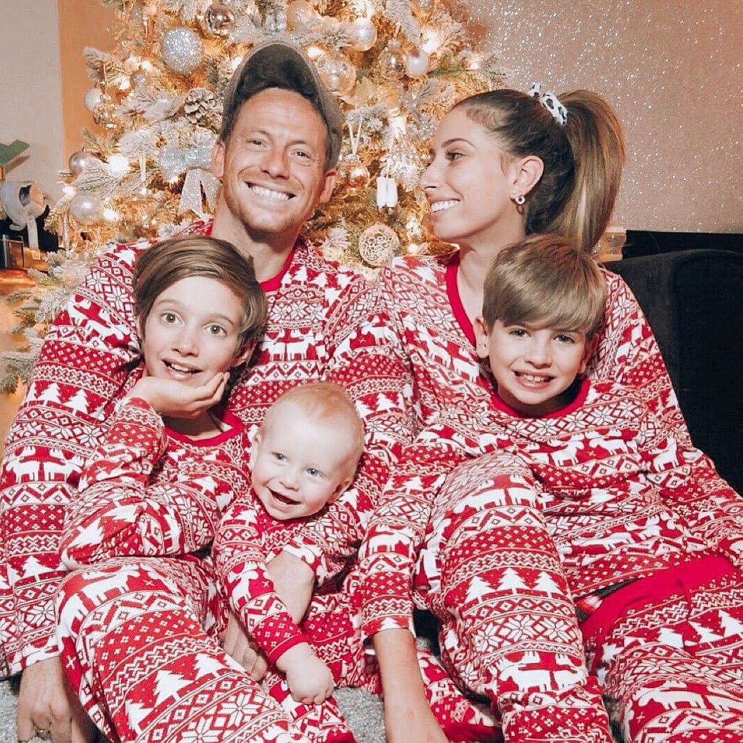 Parents and three children cuddling beside a lit tree in red Christmas pajamas with white reindeer and snowflake patterns.