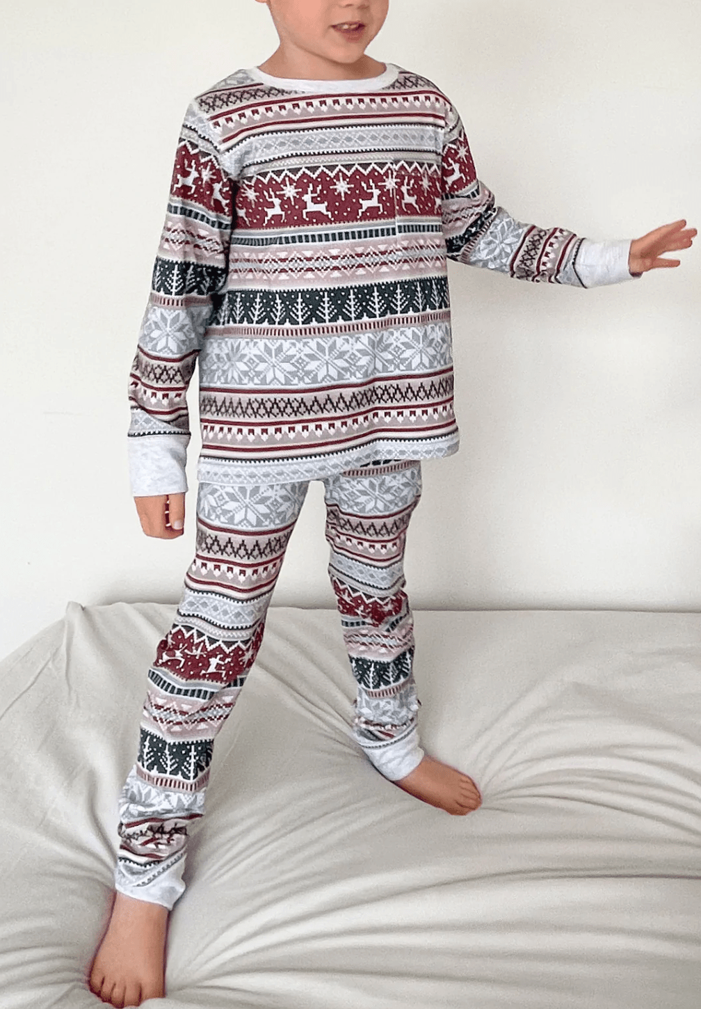 Boy Jumping on White Bed in Fair Isle Christmas Pajamas – Red Reindeer and Snowflake Stripe Print Kids Set, Playful Holiday Morning