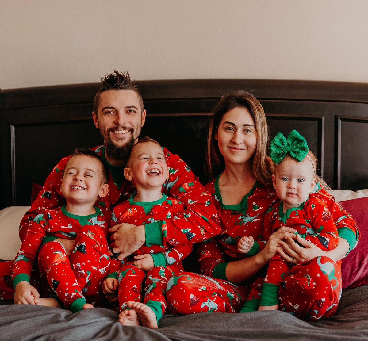 Family cuddling on a bed in red Christmas pajamas with colorful festive dinosaur prints and green cuffs.