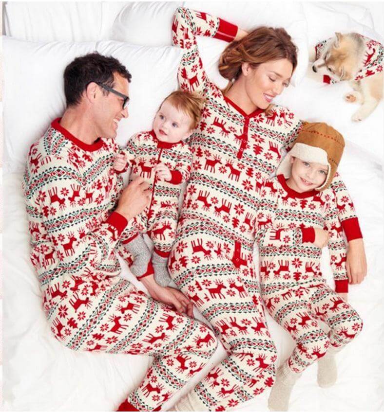 Familie liegt neben einem Hund auf einem Bett in weißen Weihnachtspyjamas mit roten Rentieren und grünen geometrischen Streifen.