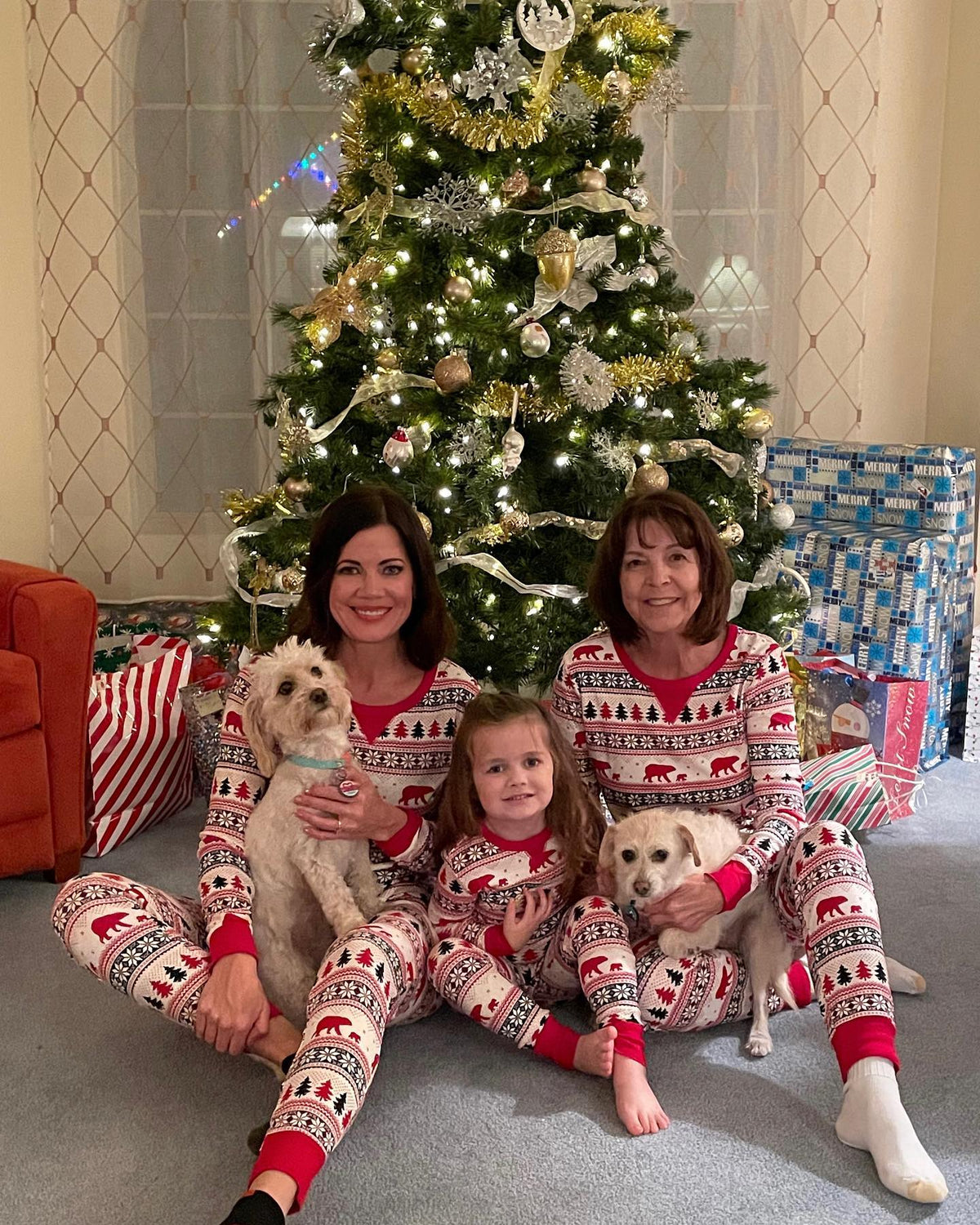 Due donne e una bambina con due cani accanto a un albero, in pigiami natalizi Fair Isle bianchi con motivi di orsi e alberi.