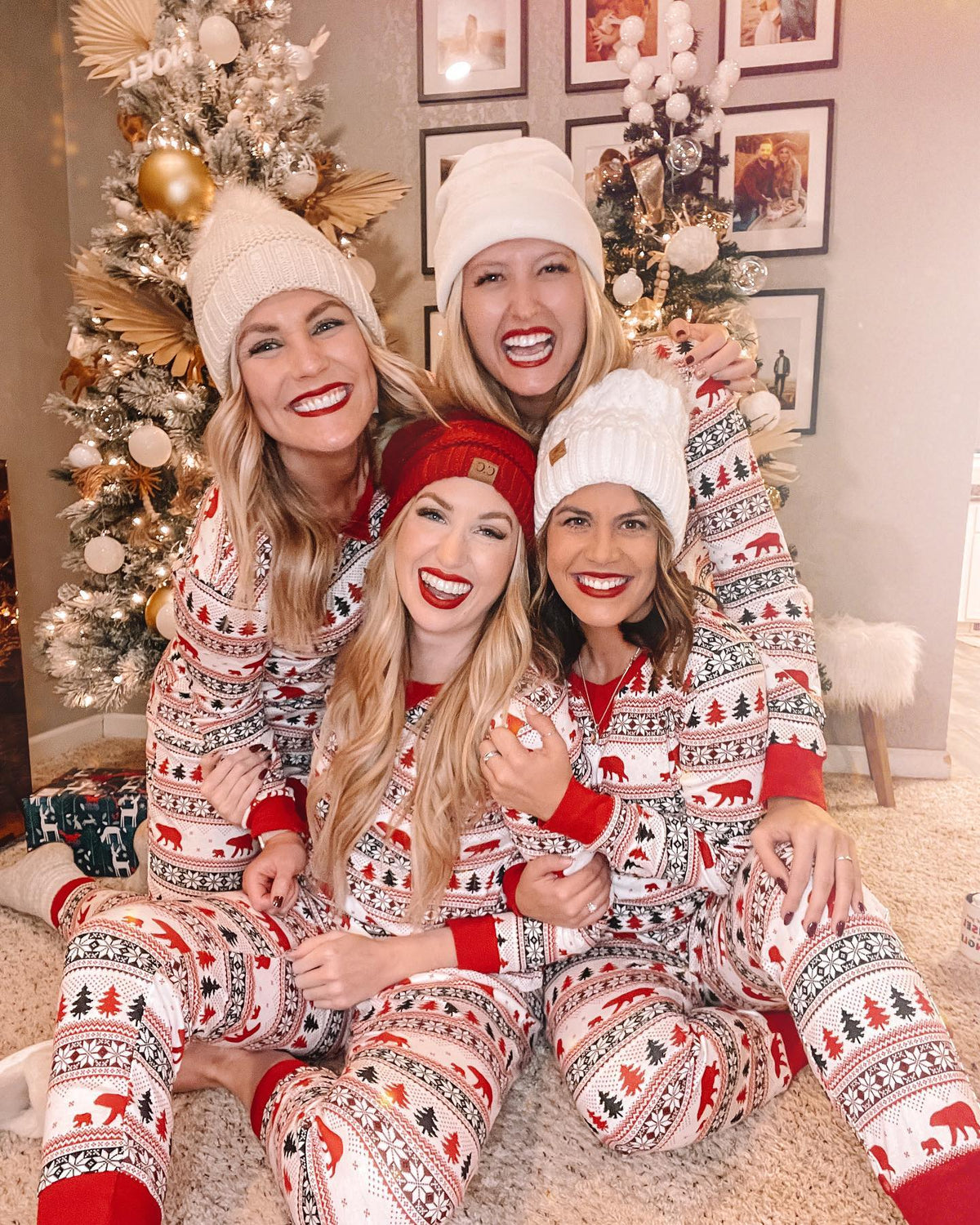 Quattro donne con berretti lavorati a maglia sedute accanto ad alberi di Natale, in pigiami Fair Isle bianchi con orsi rossi, alberi e polsini rossi.