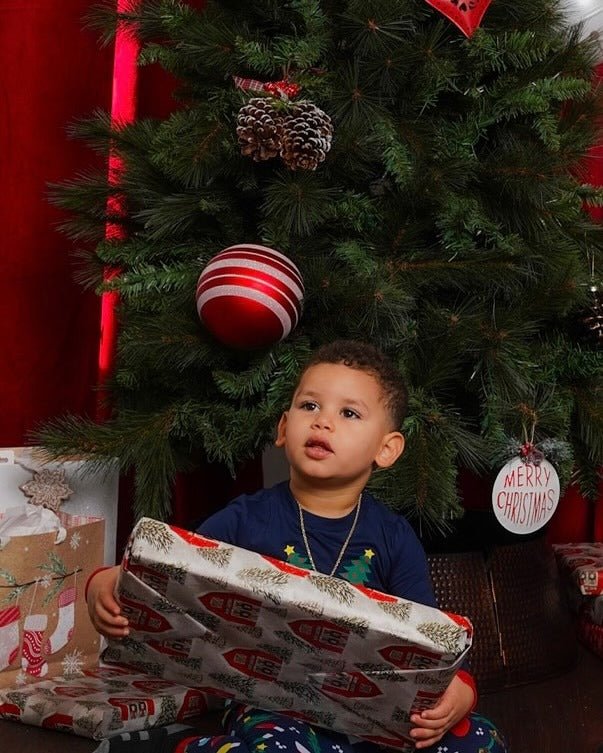 Bambino che tiene un regalo incartato sotto un albero di Natale, in pigiama natalizio blu navy con pantaloni stampati multicolori.