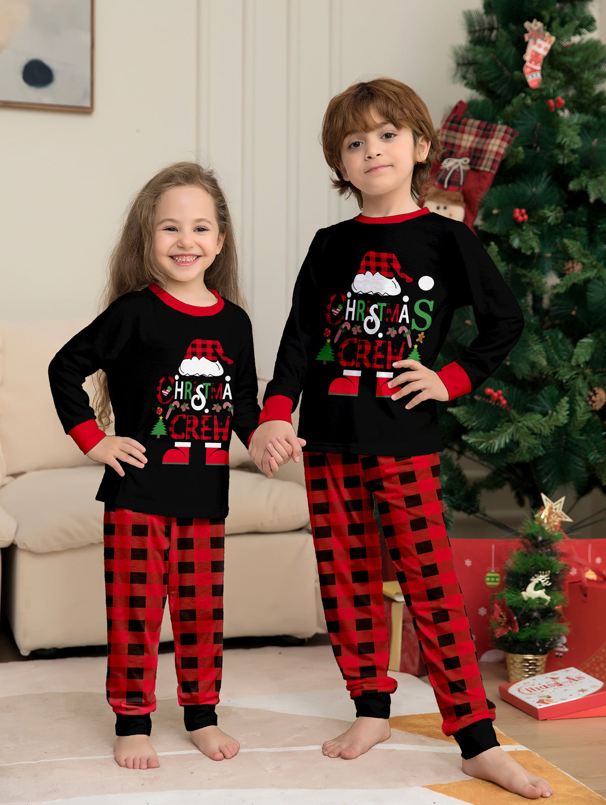 Due bambini che si tengono per mano accanto a un albero con maglie nere Christmas Crew del pigiama e pantaloni a grandi quadri rossi e neri.