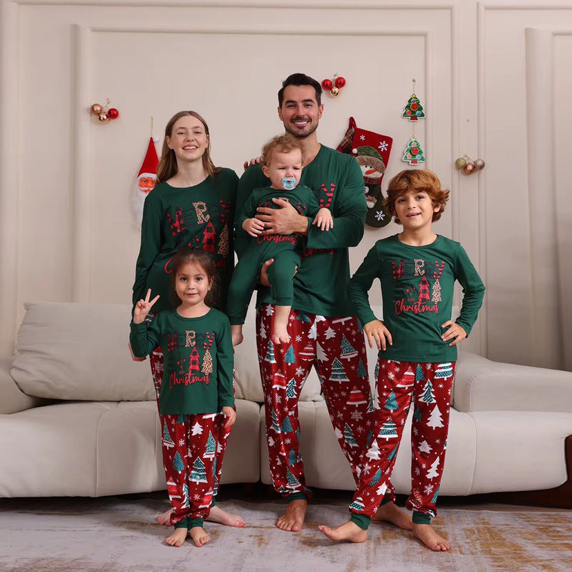 Famiglia accanto a un divano con maglie verdi Merry Christmas e pantaloni rossi del pigiama con alberi; il neonato indossa una tutina verde.