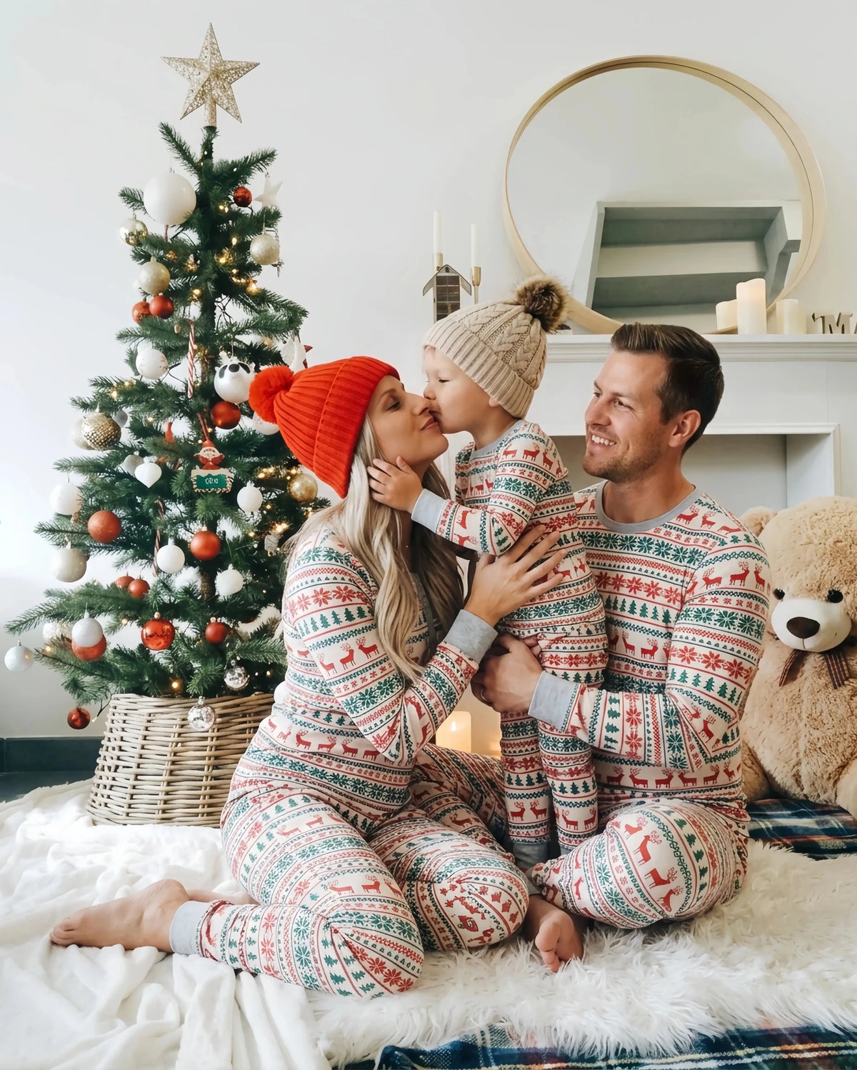 Madre che bacia un bambino tenuto dal padre accanto a un albero di Natale, tutti in pigiami coordinati con renne e alberi.