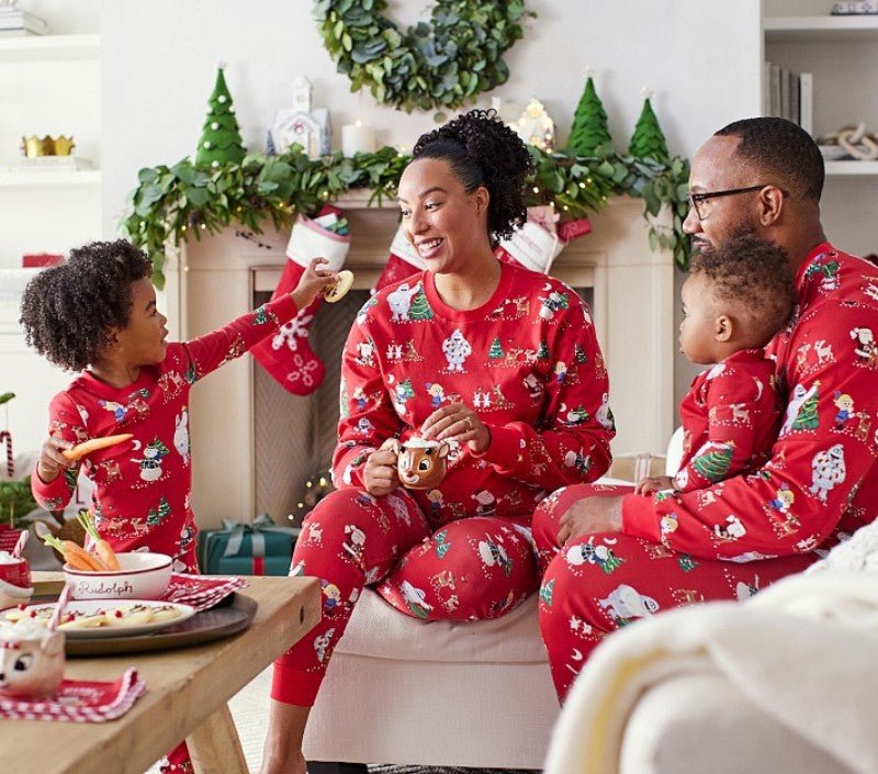 Famiglia che fa colazione accanto a un camino decorato, in pigiami natalizi rossi con pupazzi di neve, renne e alberi.