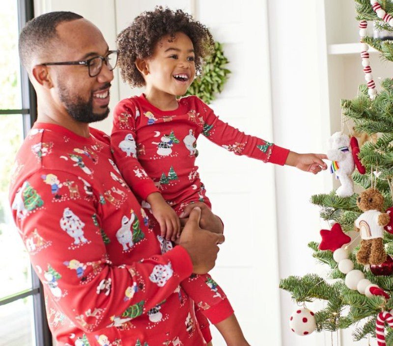 Padre che tiene un bambino sorridente accanto a un albero di Natale, in pigiami rossi coordinati con personaggi festivi.