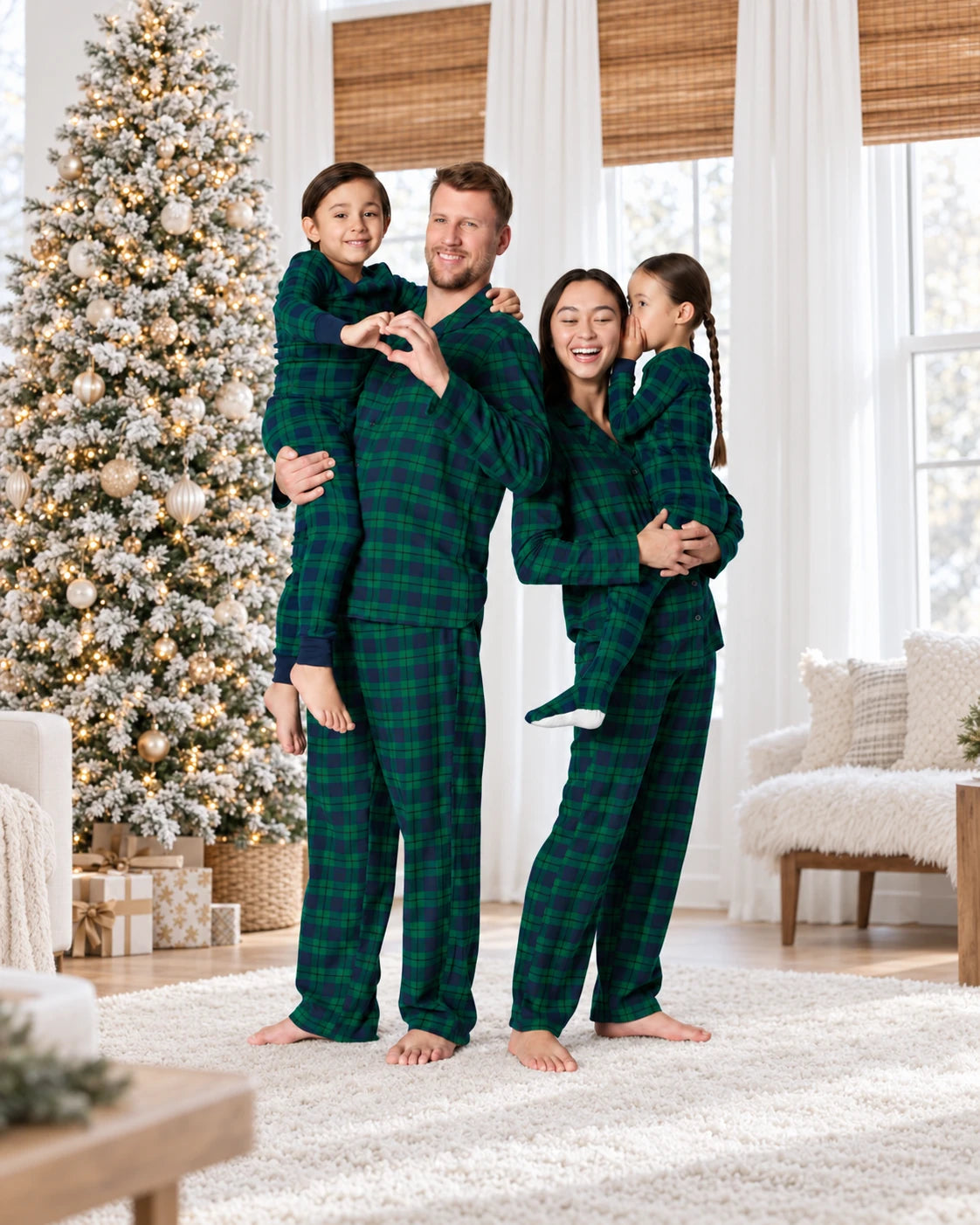 Parents holding two children in matching forest green and navy plaid pajamas beside a flocked Christmas tree.