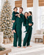 Parents holding two children in matching forest green and navy plaid pajamas beside a flocked Christmas tree.