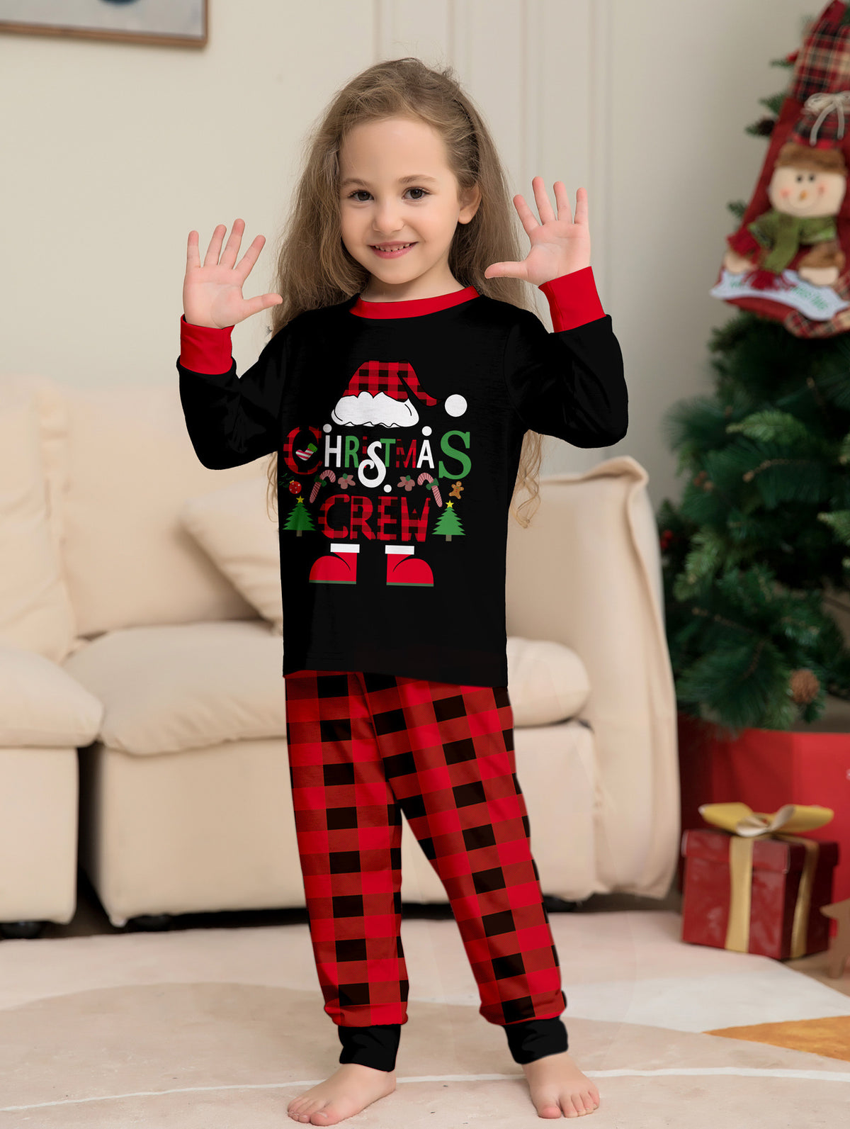 Bambina con le mani alzate, con maglia nera Christmas Crew del pigiama e pantaloni a quadri rossi e neri, accanto a un albero di Natale.