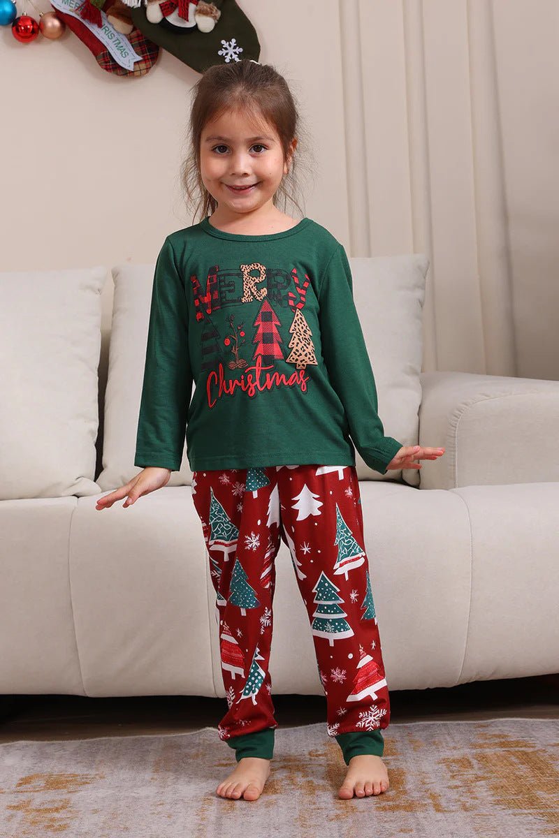 Bambina con maglia verde Merry Christmas del pigiama e pantaloni rossi con alberi e bordi verdi, accanto a un divano.