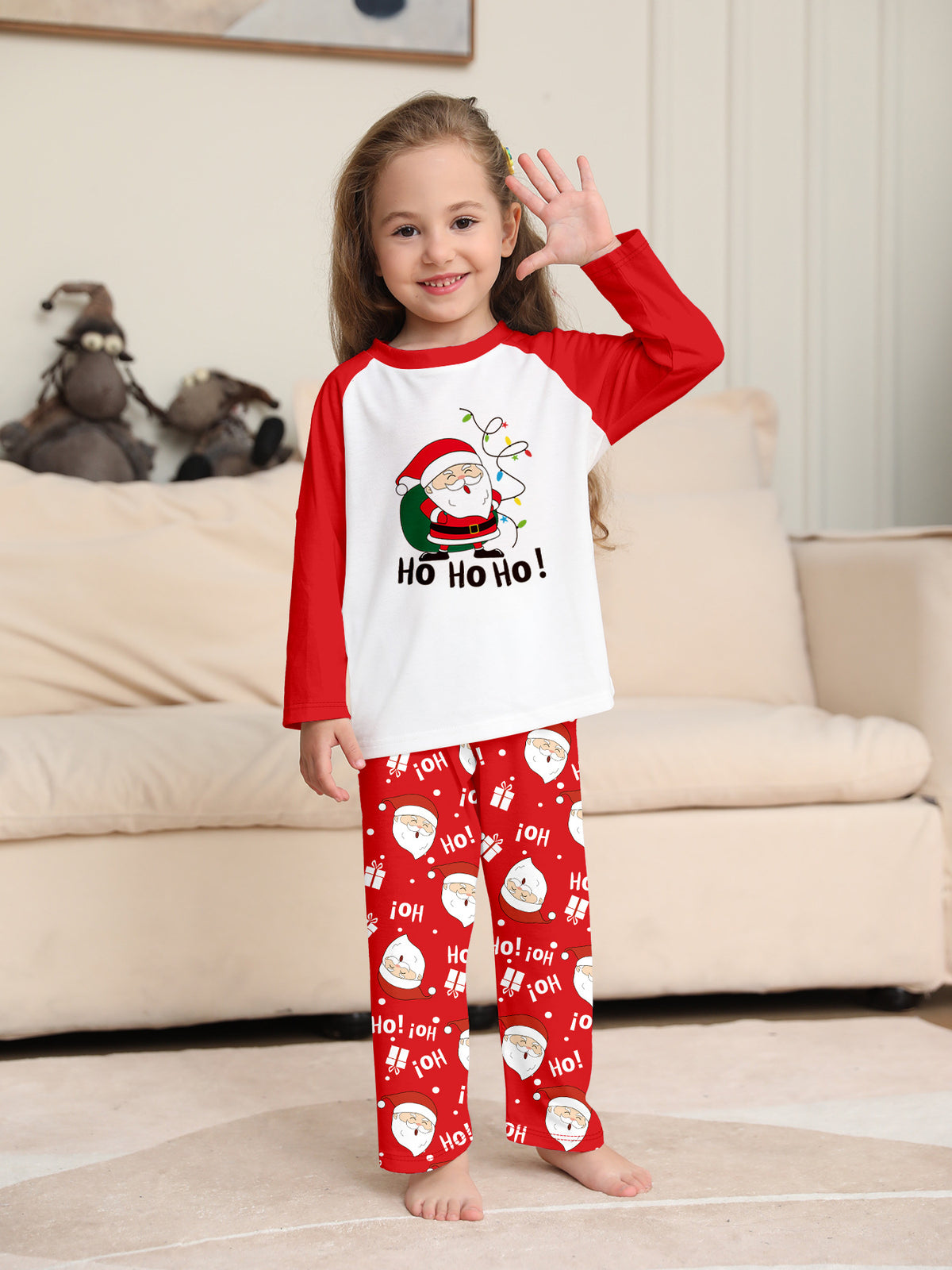 Girl waving in a white Ho Ho Ho Santa pajama top with red sleeves and red Santa print pants beside a sofa.