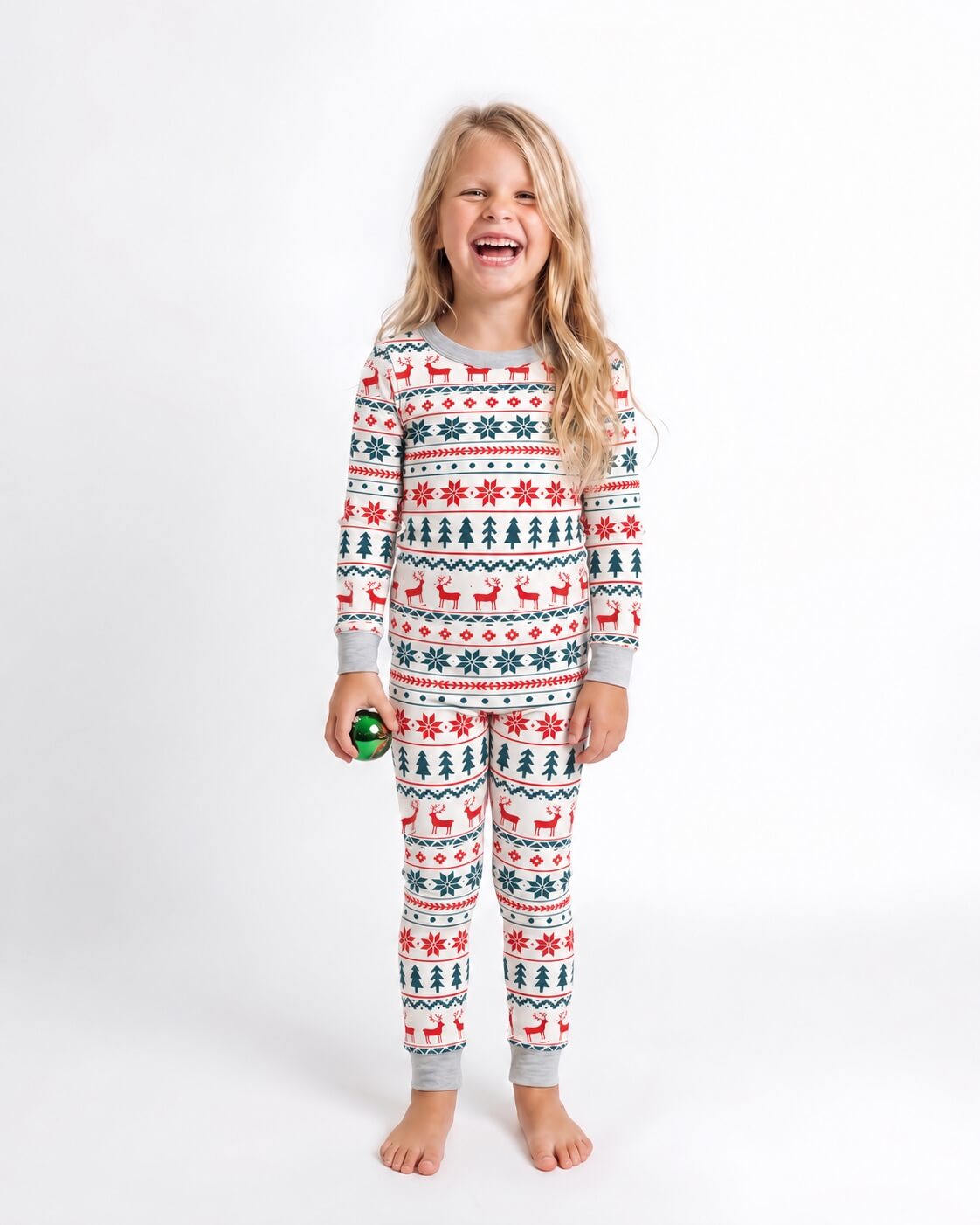 Bambina con una decorazione verde in mano, in pigiama natalizio bianco con renne rosse, alberi verdi e polsini grigi.