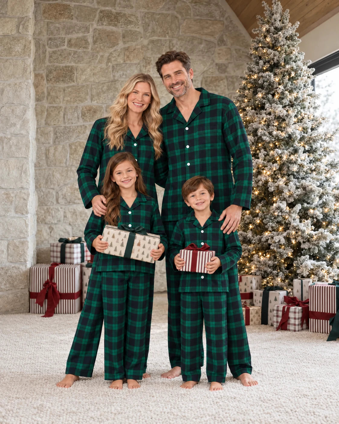 Family in green and black plaid pajamas holding presents beside a frosted tree and stone wall.