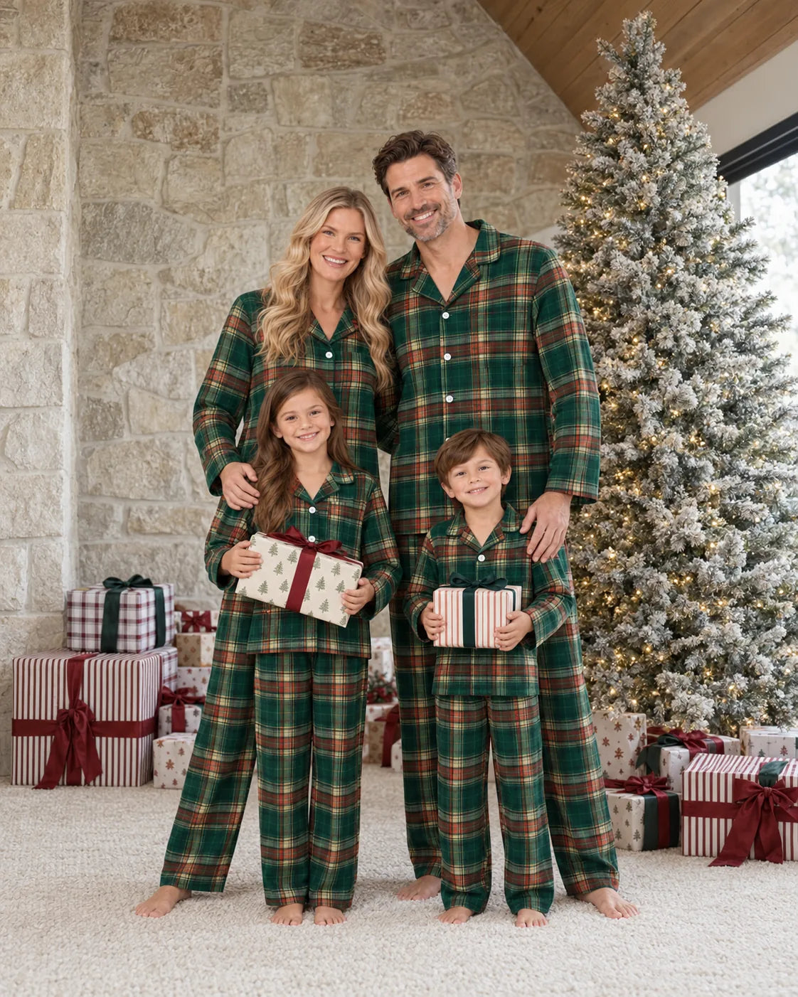 Family in light green tartan pajamas holding presents beside a frosted Christmas tree and stone wall.