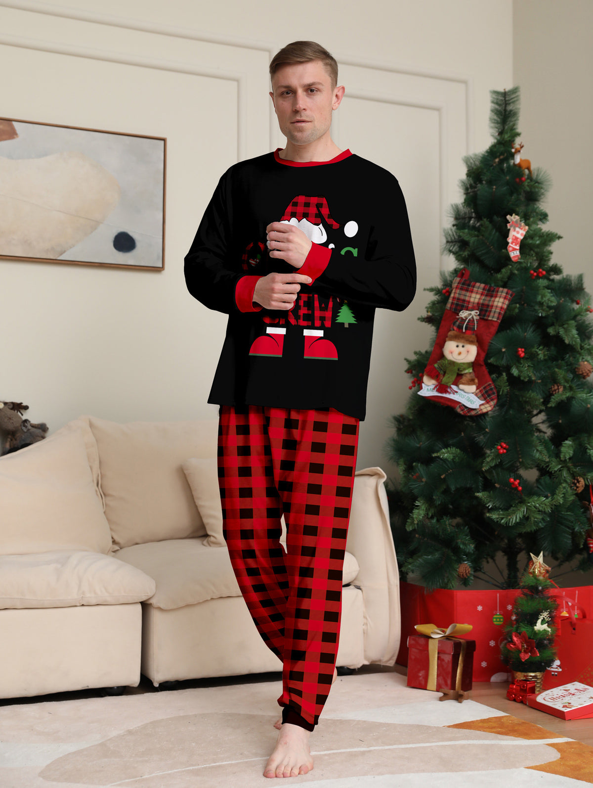 Uomo in posa accanto a un albero di Natale con maglia nera Christmas Crew del pigiama e pantaloni a grandi quadri rossi e neri.
