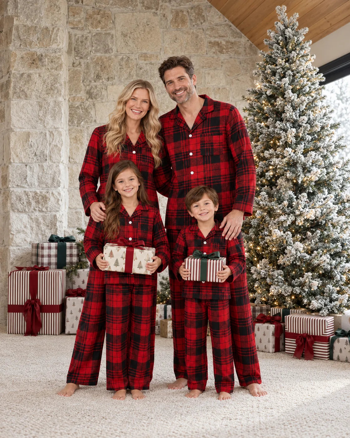Family of four in red and black tartan pajamas with gifts beside a flocked tree and stone wall.